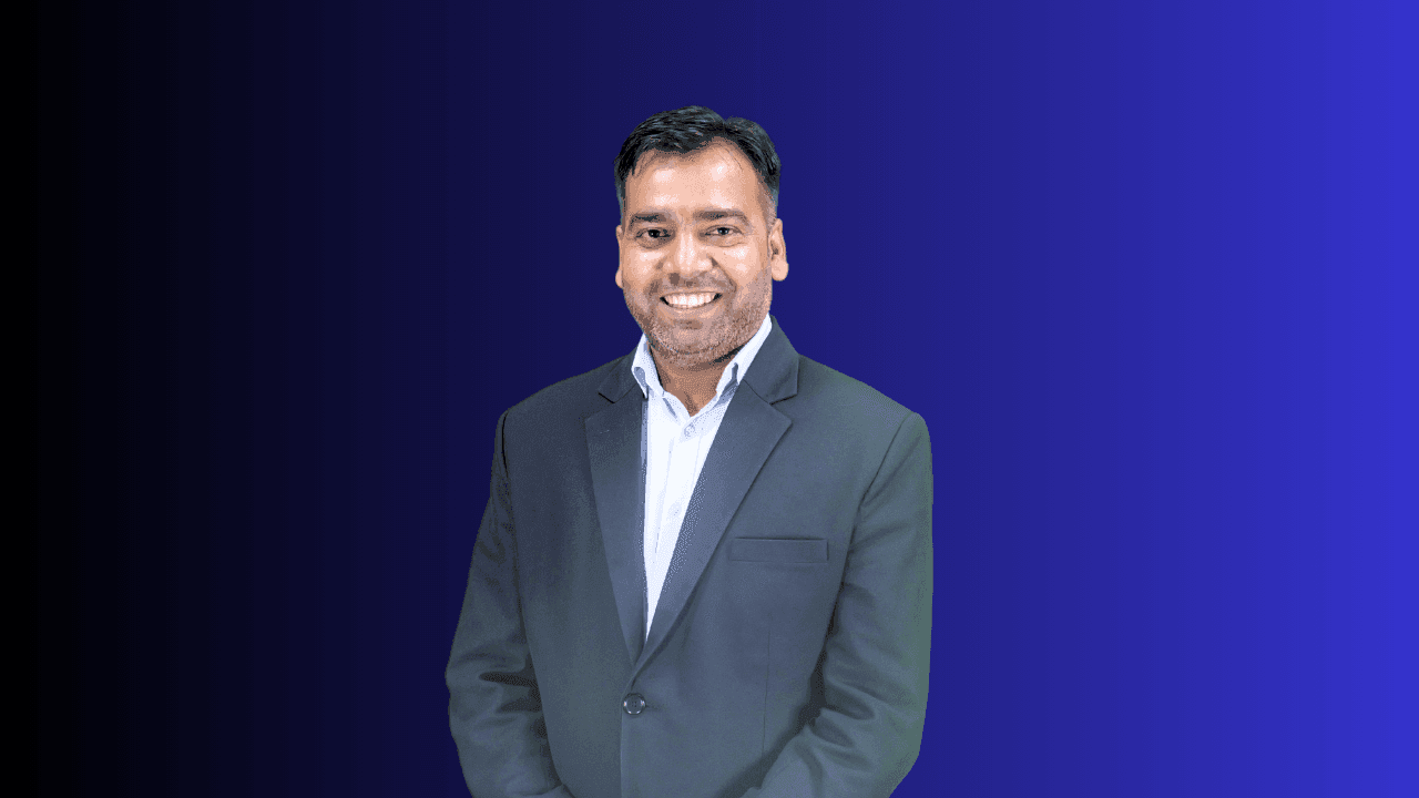 Man in formal attire wearing a dark suit and white shirt, standing against a blue gradient background.