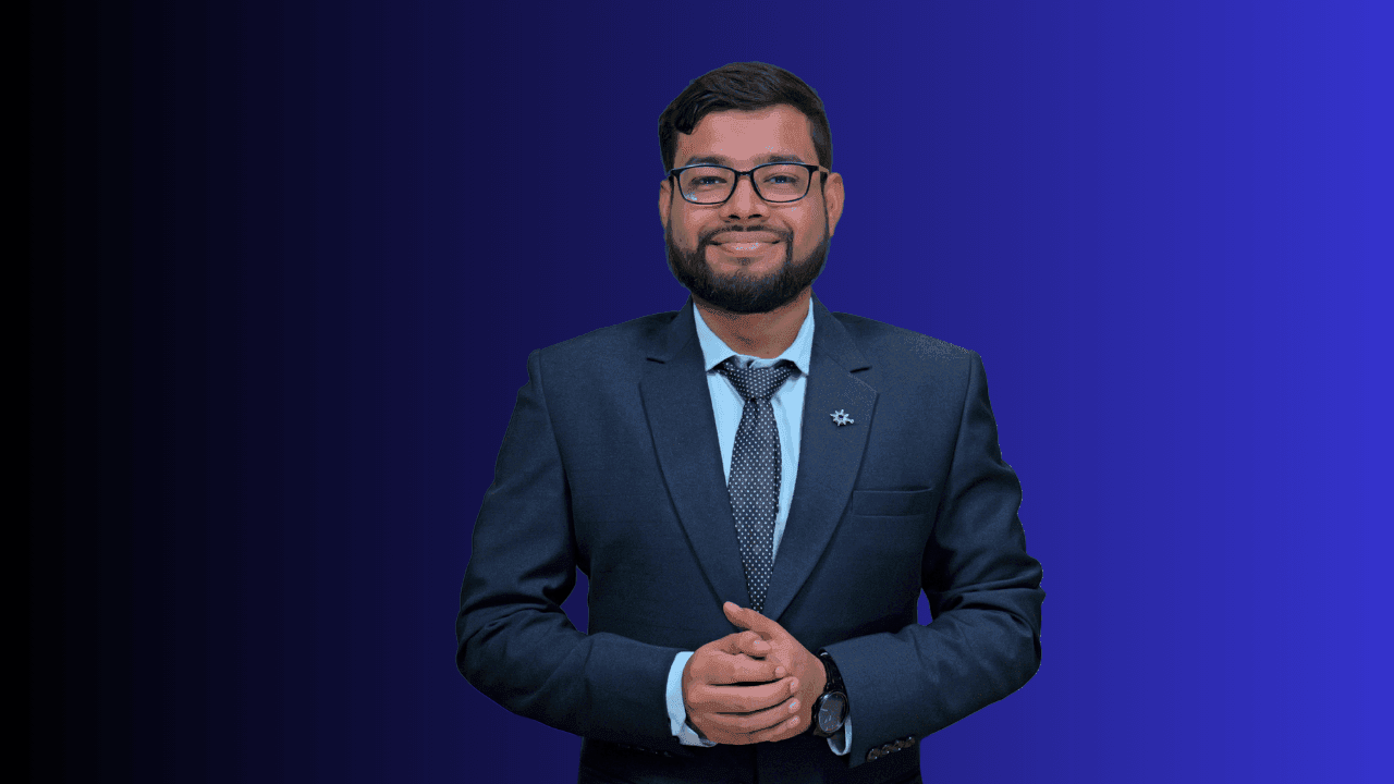 Smiling man in a dark suit and tie, standing against a gradient blue background.